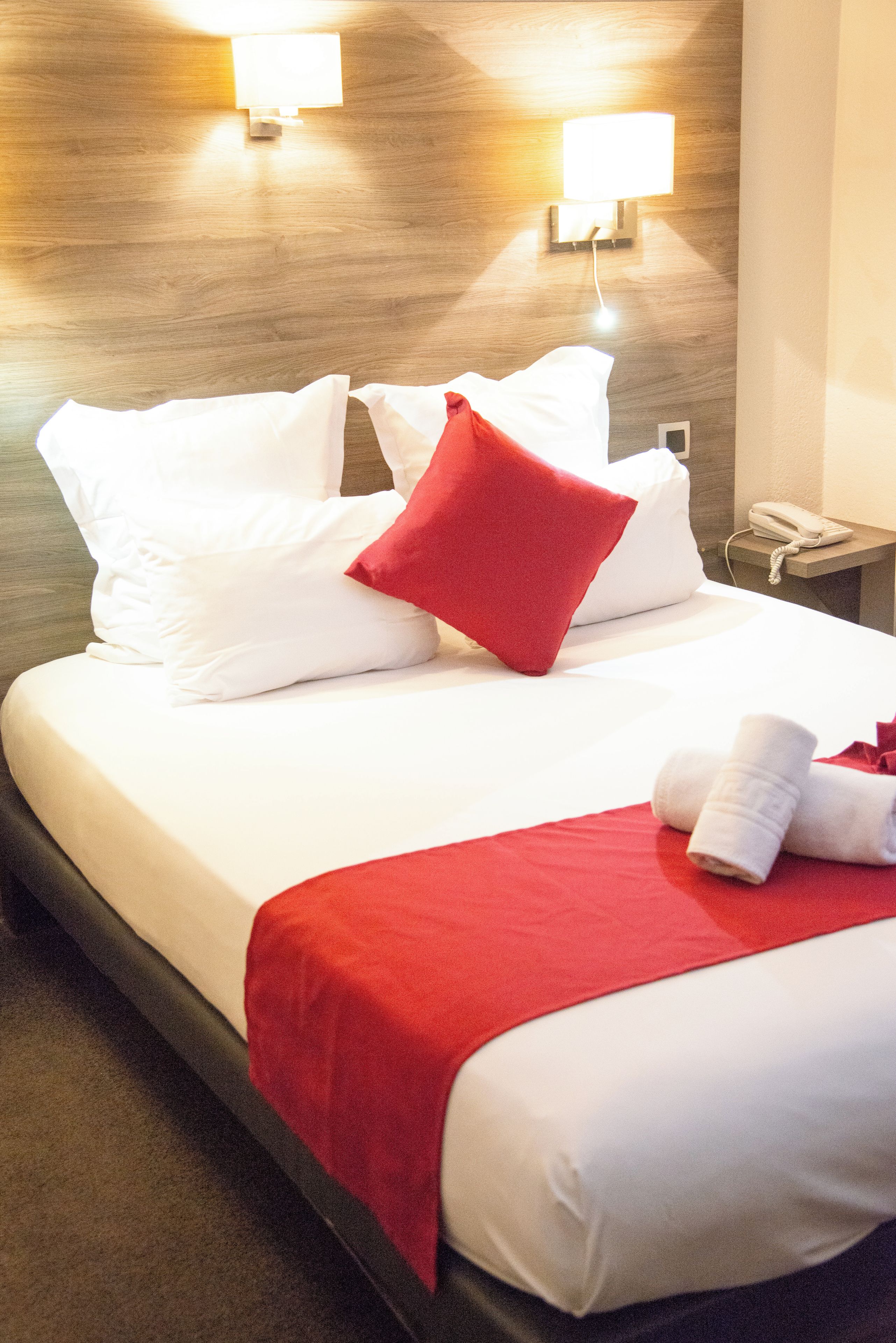 A hotel room with a bed, white bedding, a red throw blanket, and a telephone on a bedside table.