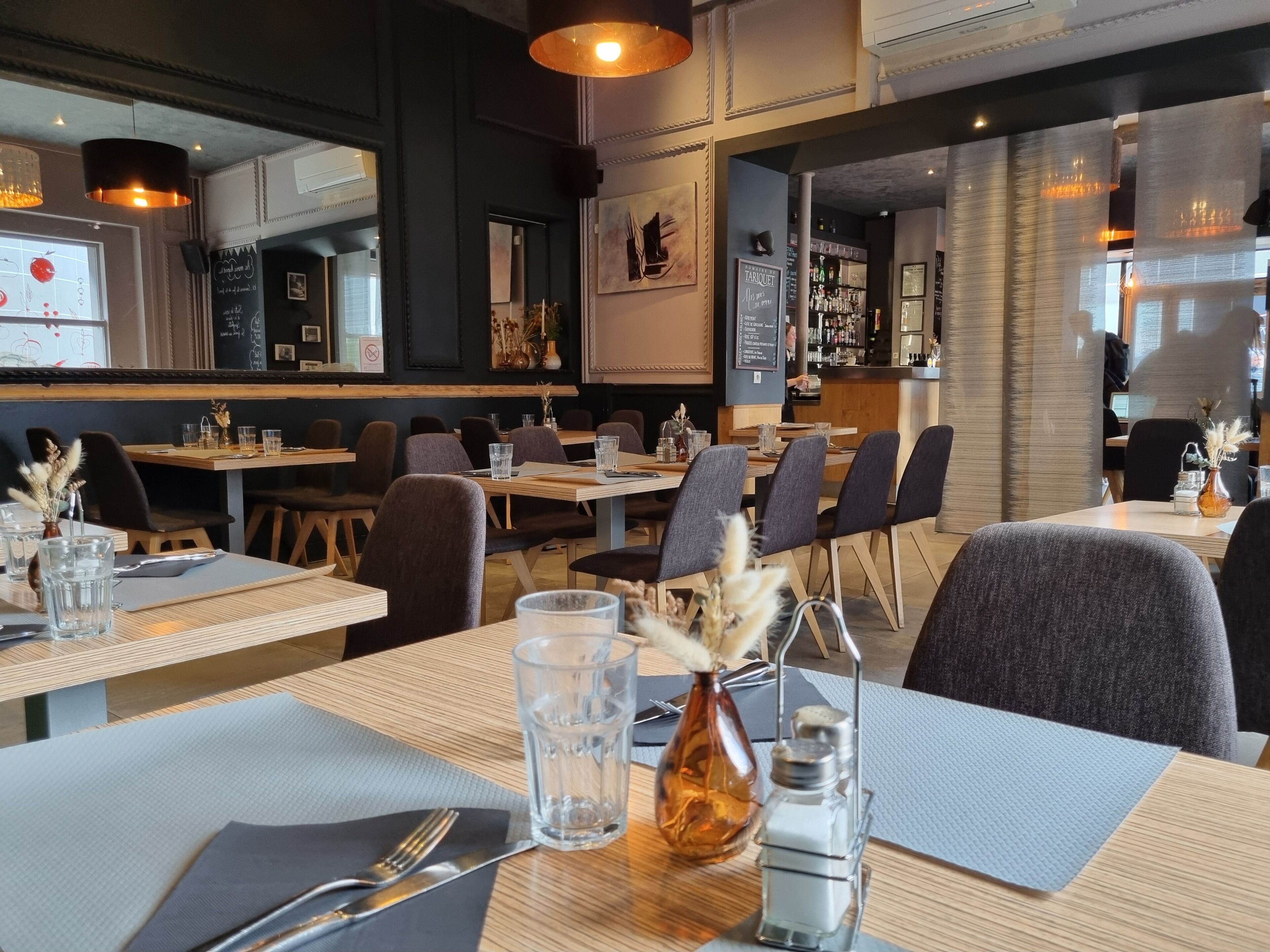 L’intérieur d’un restaurant, avec des tables dressées pour le service, un bar et une vue sur la rue.