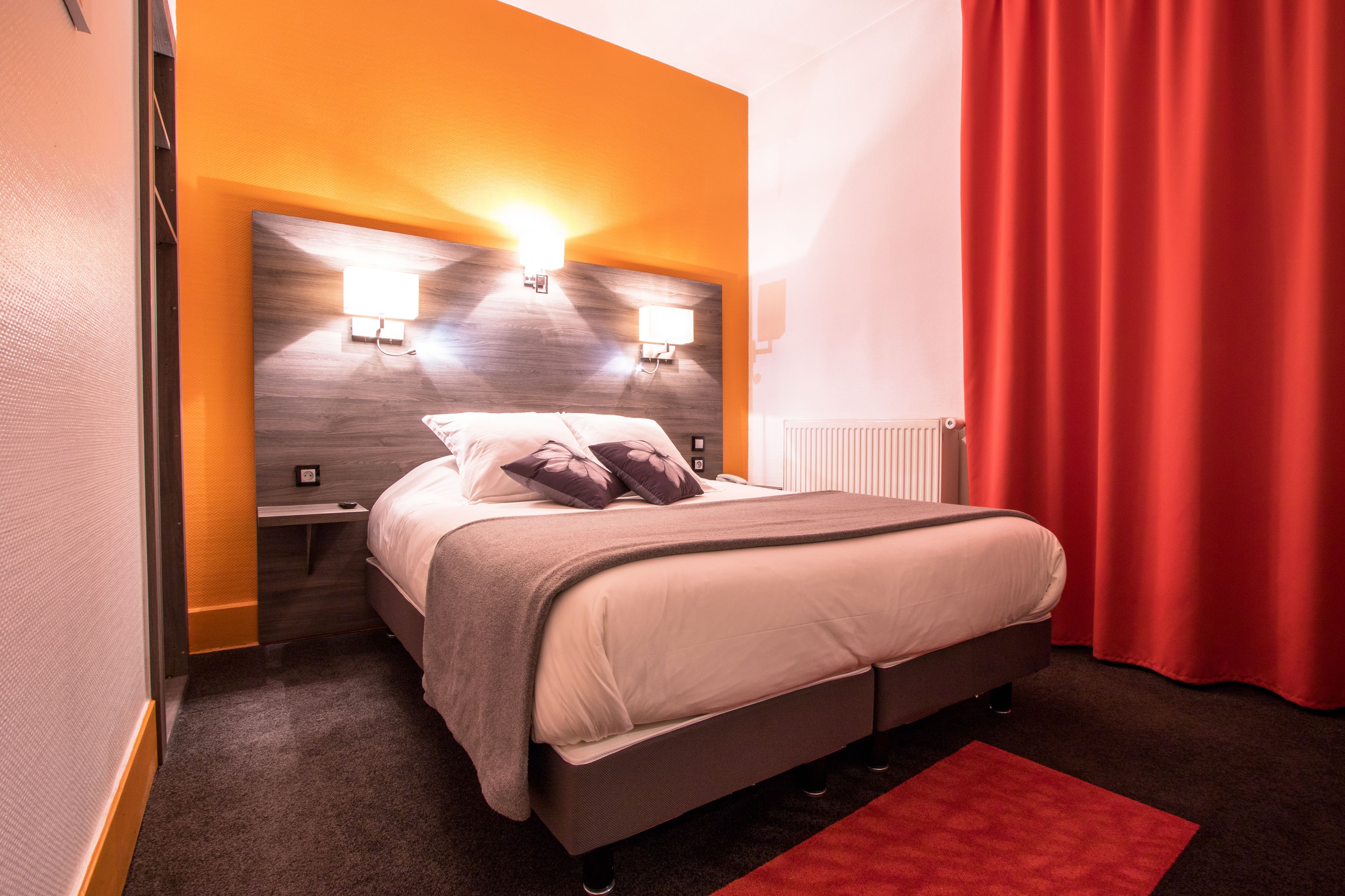 A modern hotel room with a bed, two bedside lamps, a red curtain, and a wooden headboard.