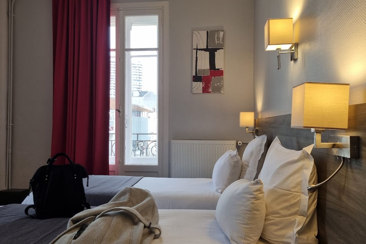 Quarto de hotel com uma cama, janela com cortinas vermelhas, radiador e luminárias fixadas na parede.