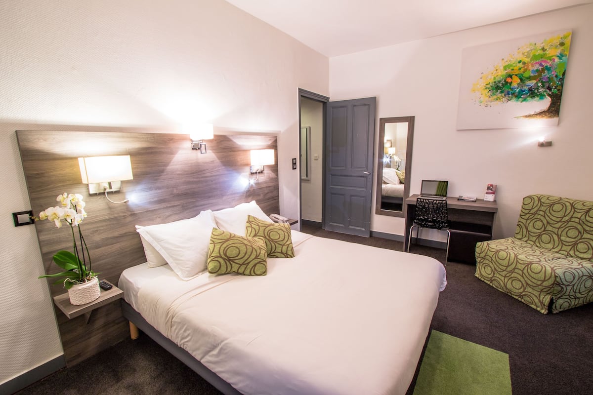 Quarto de hotel com cama, mesa de cabeceira, luminária, cadeira, escrivaninha e um quadro na parede.