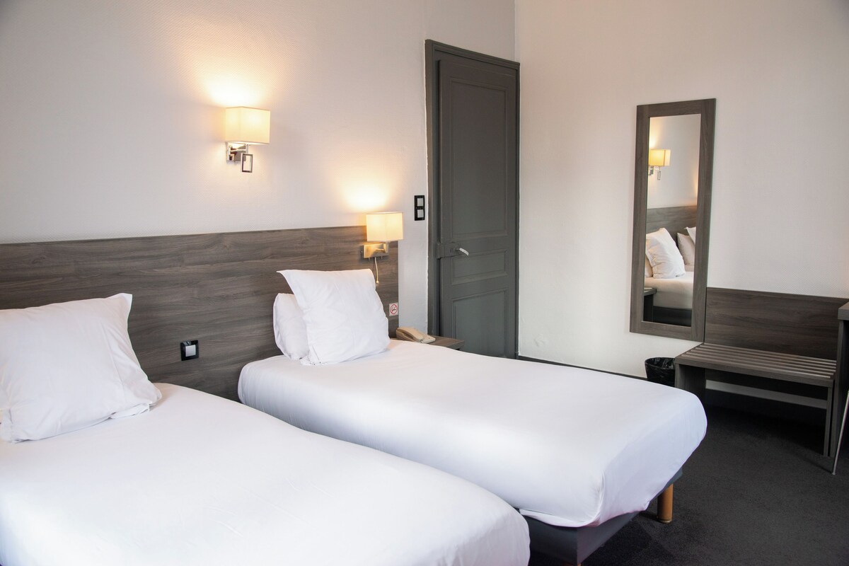 A hotel room with two single beds, a wooden headboard, bedside lamps, a mirror, and a desk.
