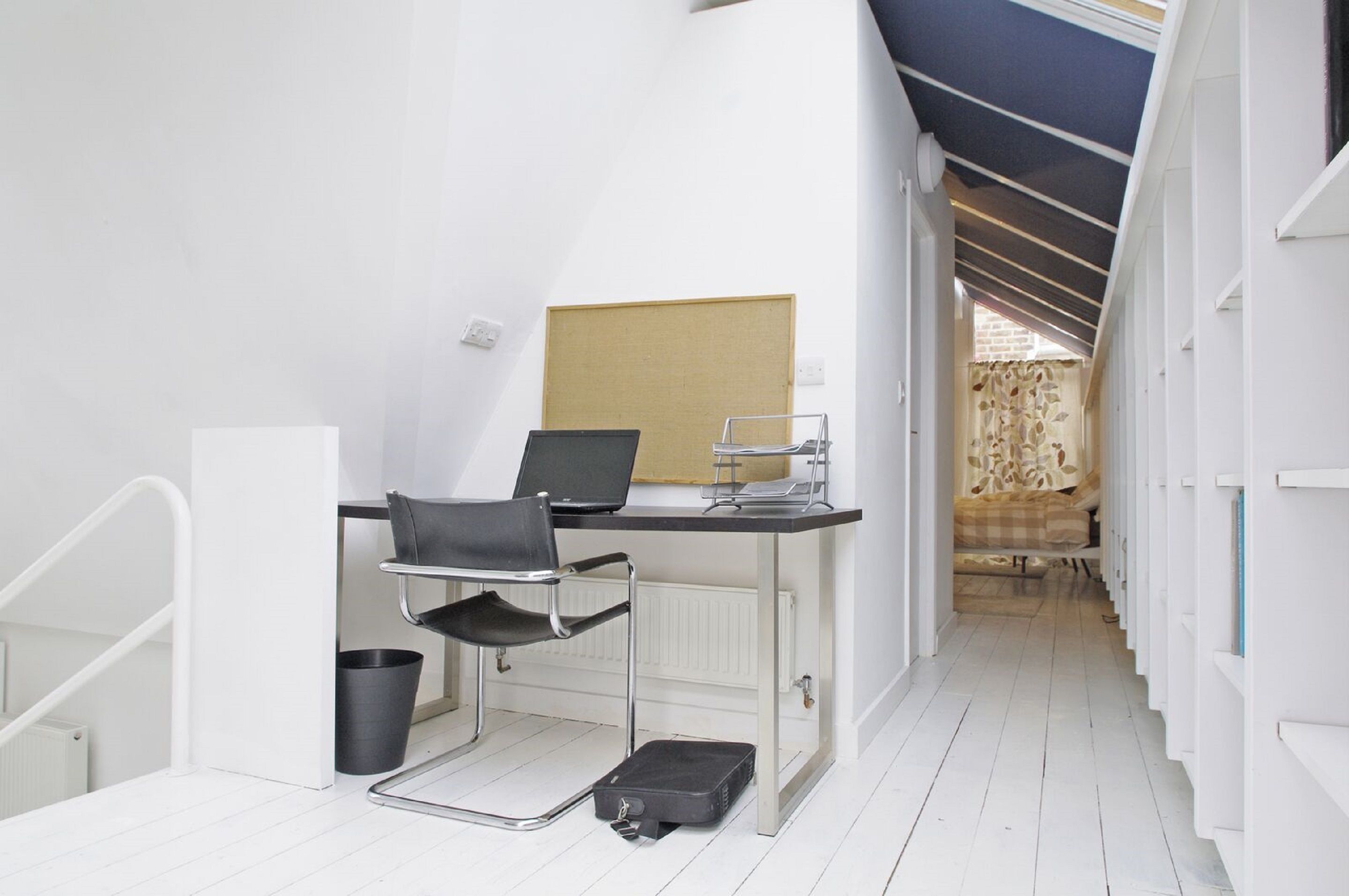 Una habitación compacta y moderna con un escritorio blanco, una silla negra y una computadora portátil. Un radiador se encuentra debajo del escritorio, y una maleta negra está en el suelo.