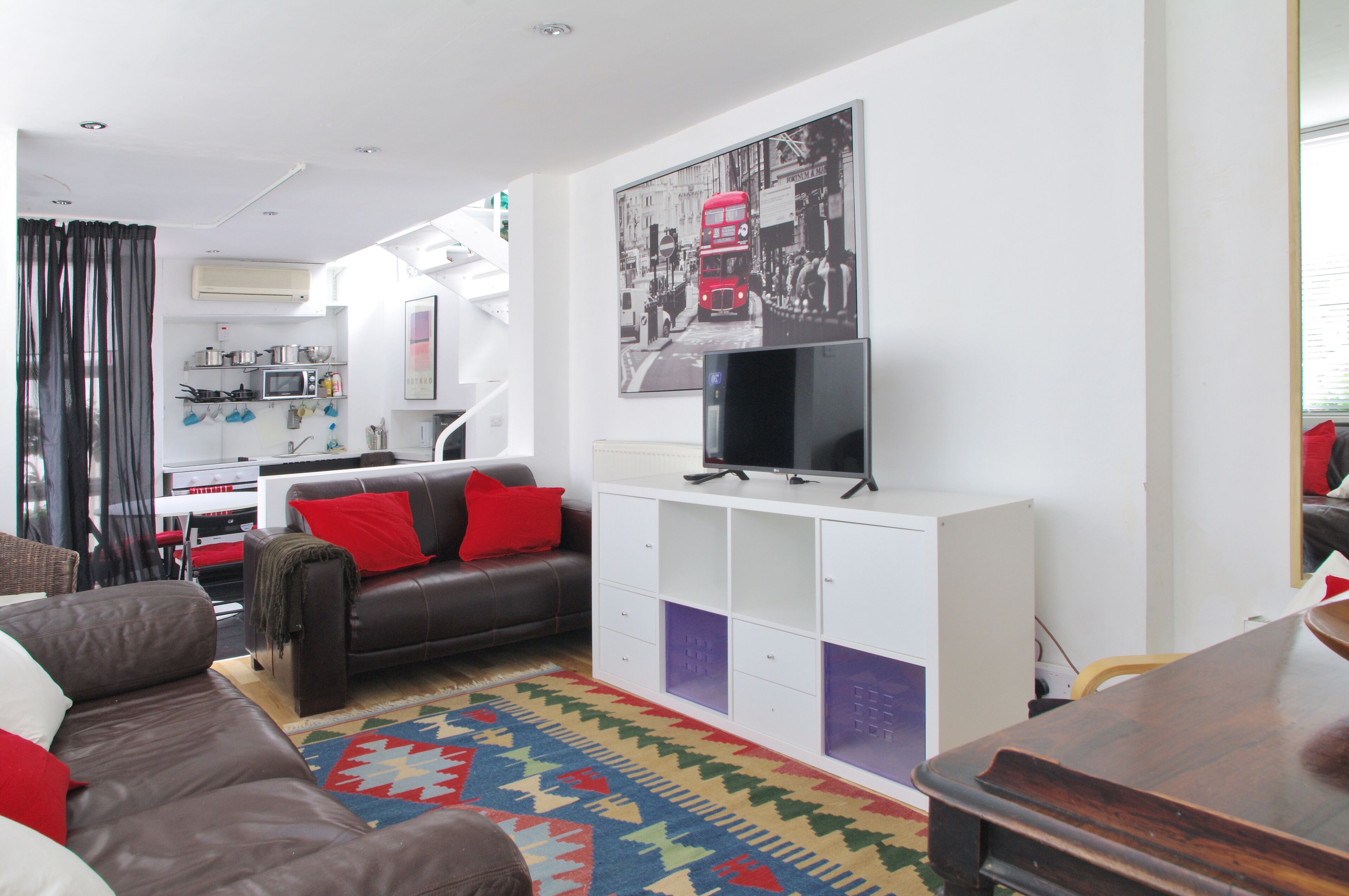 Un salon moderne avec un canapé en cuir marron, un tapis coloré, un téléviseur à écran plat posé sur un meuble blanc et un escalier menant à un niveau supérieur.