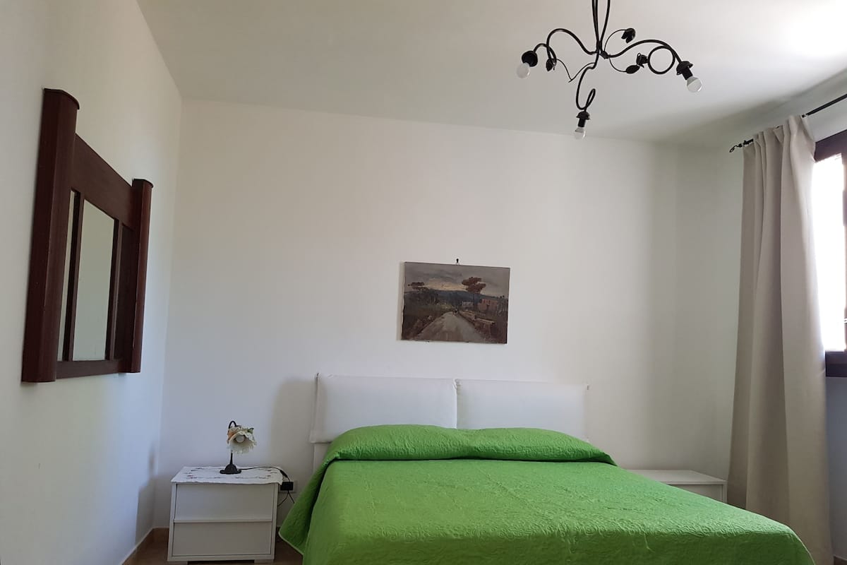 A bedroom with a bed, a nightstand, a painting on the wall, and a window with curtains.