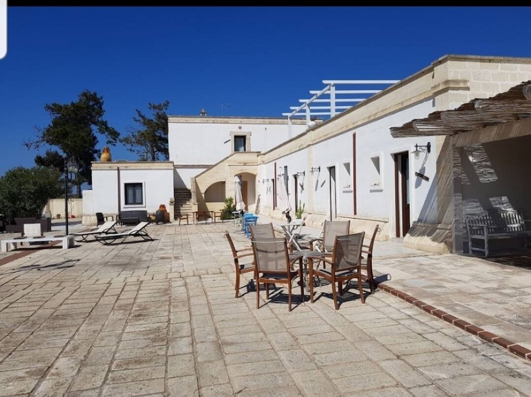 Ein Außenbereich mit Terrasse, Tisch und Stühlen, einem überdachten Sitzbereich und einem Gebäude mit weißer Fassade und Balkon.