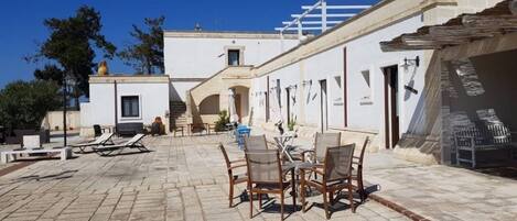 Ein Außenbereich mit Terrasse, Tisch und Stühlen, einem überdachten Sitzbereich und einem Gebäude mit weißer Fassade und Balkon.