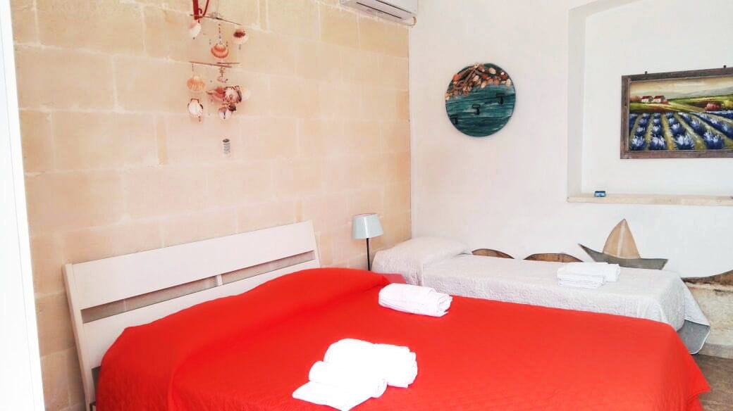 A bedroom with a red bedspread, a white headboard, a wall-mounted wind chime, a round wall decoration, and a framed painting.