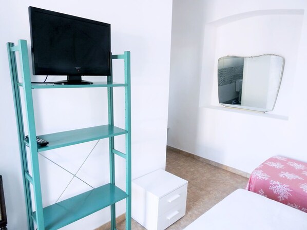 A room with a teal shelving unit holding a TV, a small white drawer, a bed with a pink patterned bedspread, and a wall-mounted mirror.