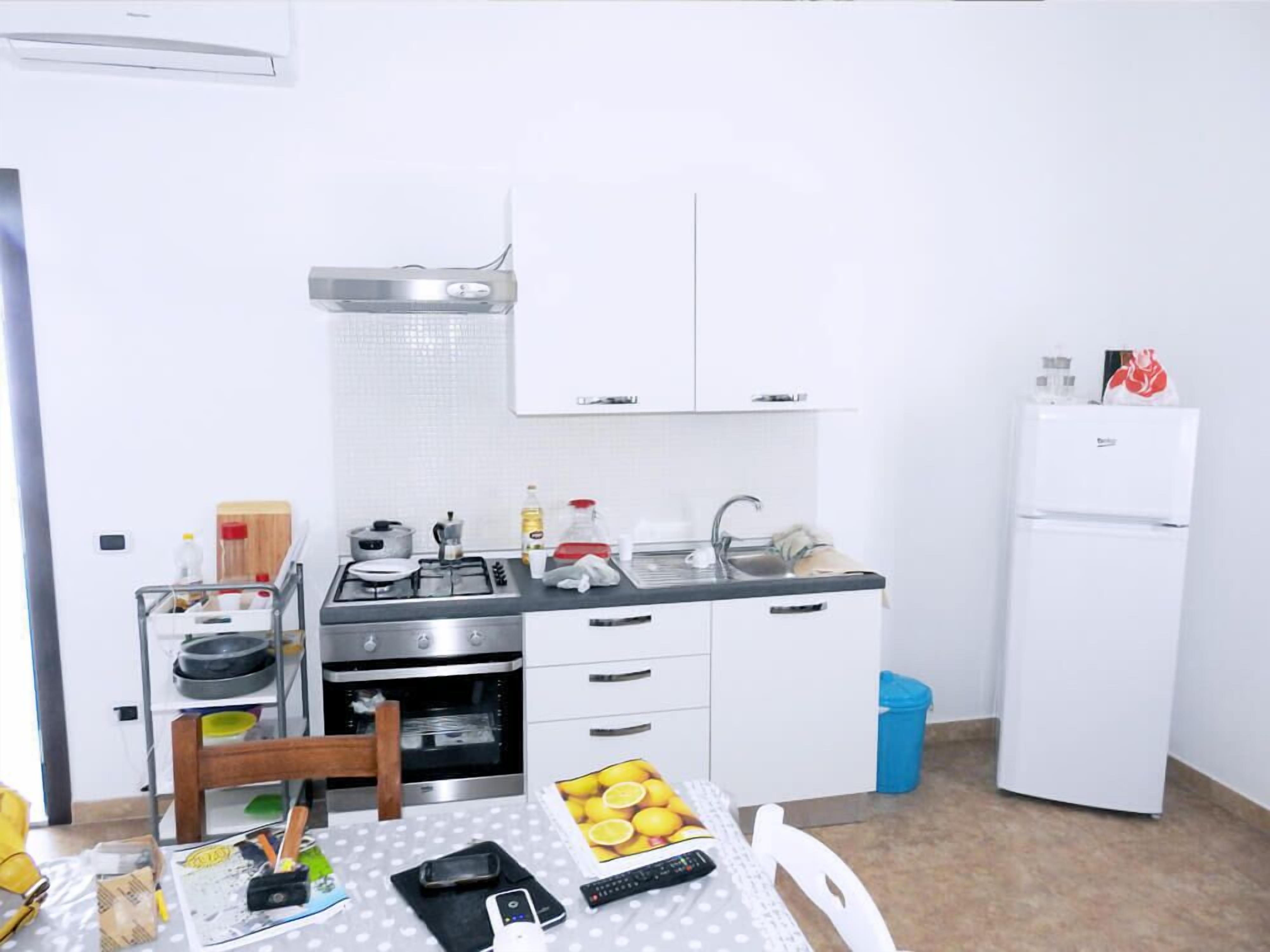 A small kitchenette with a stove, sink, and white cabinets. There is a table with a polka dot tablecloth and a chair.