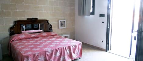 A bedroom with a bed, a wooden headboard, a brick wall, a door with a curtain, and a small bedside table with a picture.