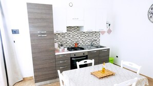 A compact kitchen with a built-in stove, a sink, and a small oven. There is a dining table with a patterned tablecloth and white chairs. - Masseria Prisuti (Manduria)