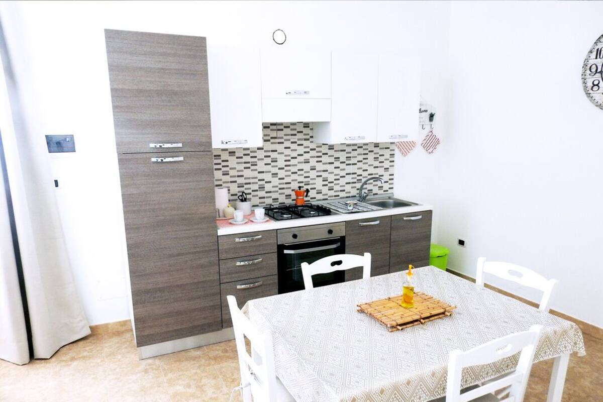 A compact kitchen with a built-in stove, a sink, and a small oven. There is a dining table with a patterned tablecloth and white chairs.