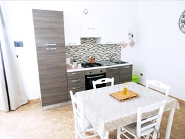 Una cocina compacta con estufa integrada, un lavamanos y un horno pequeño. Hay una mesa de comedor con mantel estampado y sillas blancas.