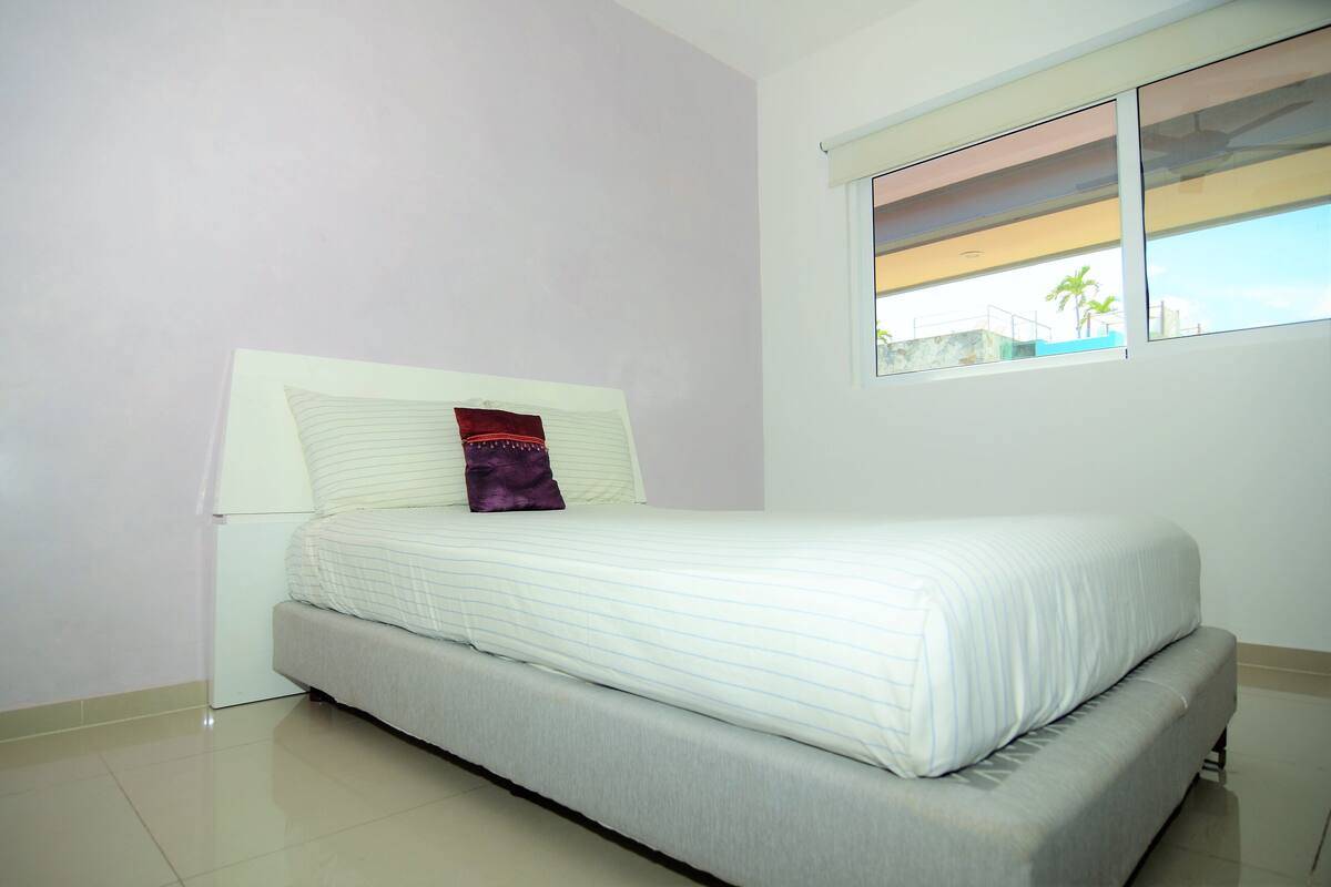 A neatly made bed with a white headboard and a single purple pillow, a window with a view of palm trees, and a white wall.