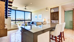 A modern kitchen with a large island, a dining area with bar stools, a sofa, and a view of the city through large windows. - Carmen Sol Rentals Apartamentos (Playa del Carmen)