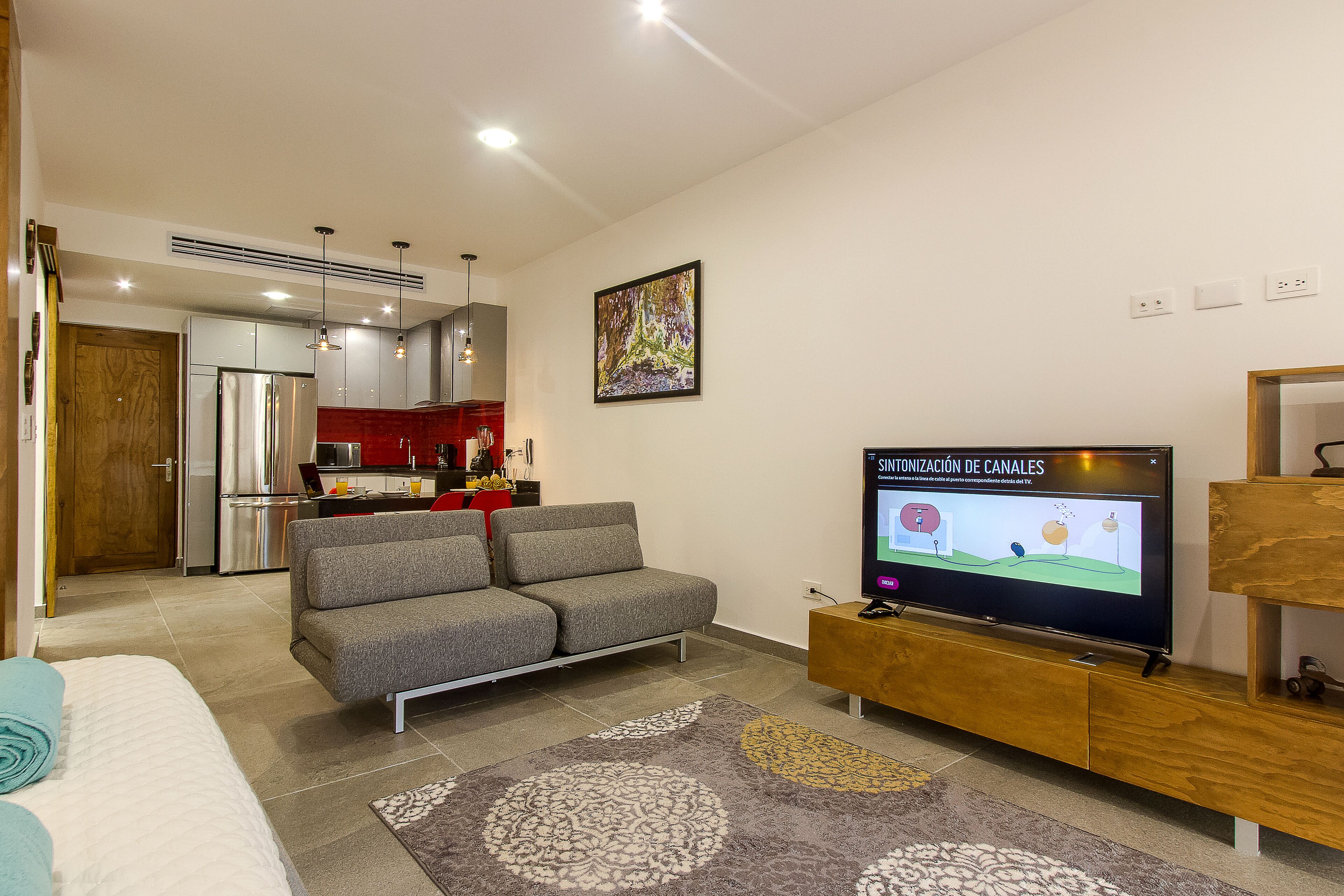 A modern living room with a grey sofa, a TV on a wooden stand, and a kitchen in the background.