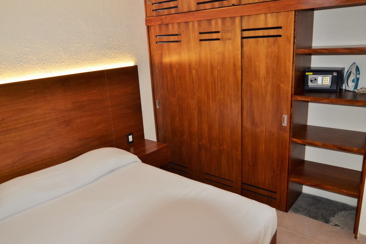 A bedroom with a wooden headboard, a bed with white bedding, a wooden wardrobe with a microwave on top, and a shelf with a kettle.