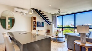 A modern living room with a dining area, a staircase, a large window with a city view, and a flat-screen TV mounted on the wall. - Carmen Sol Rentals Apartamentos (Playa del Carmen)
