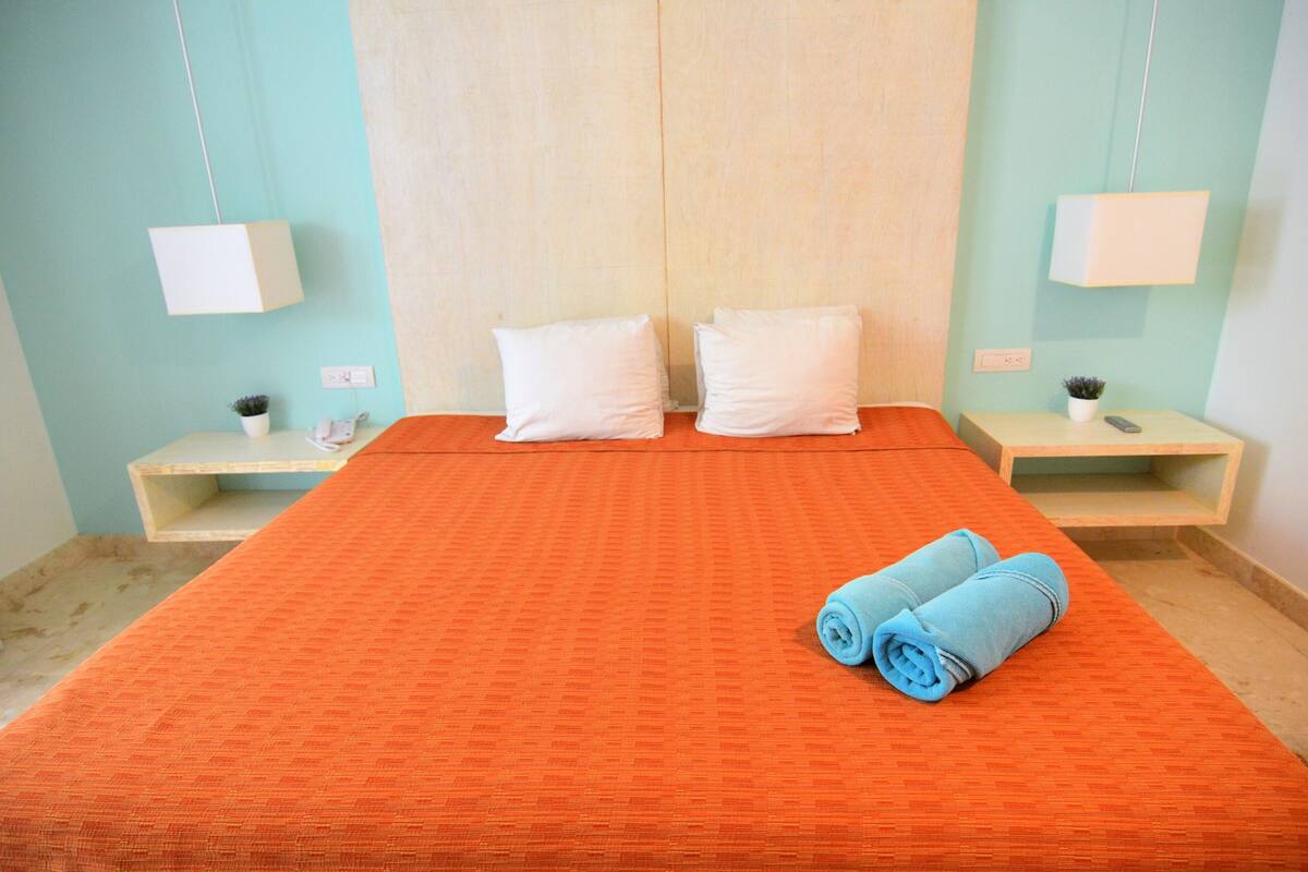 A hotel room with a large bed, two bedside tables, a nightstand, a wall-mounted lamp, and a small potted plant.