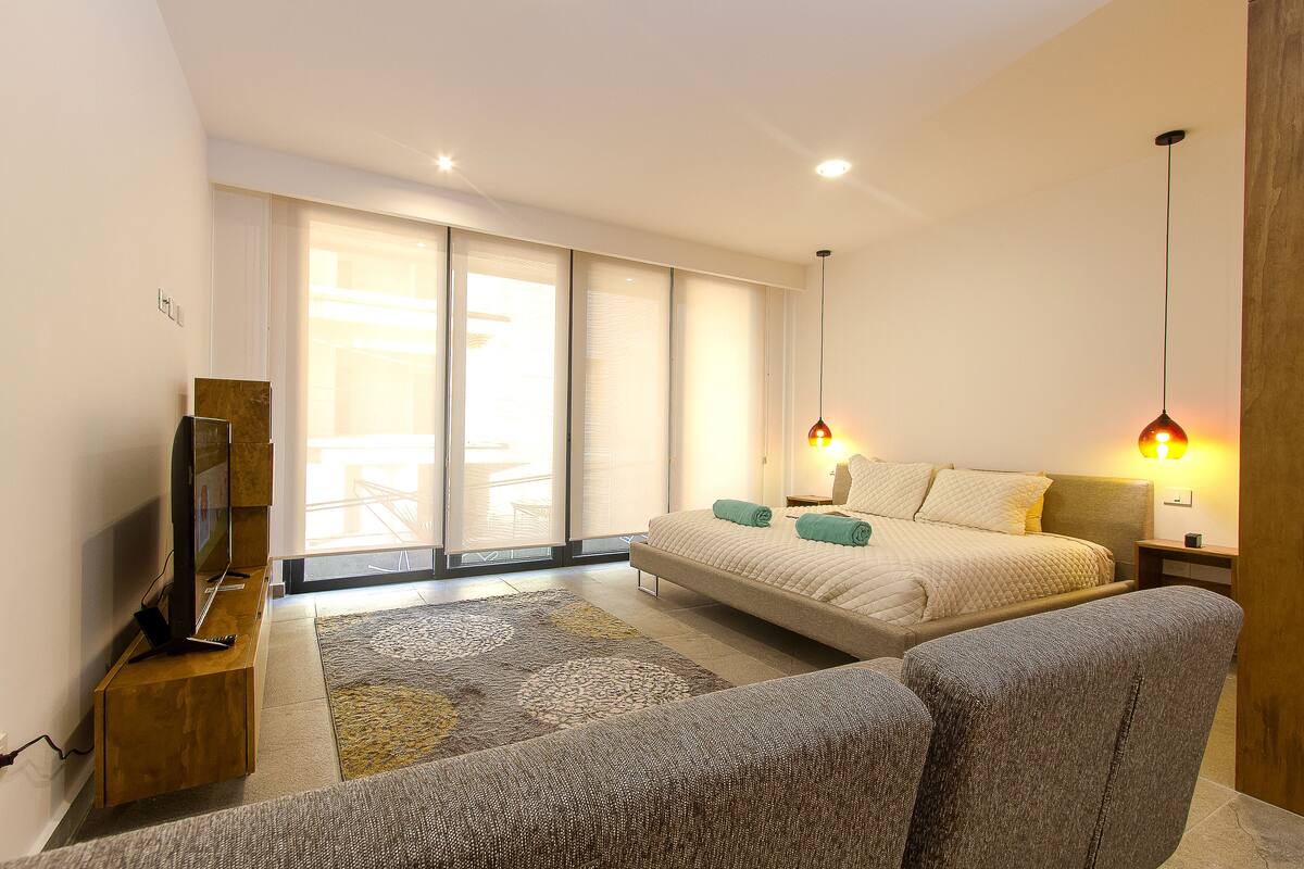 A modern bedroom with a bed, a TV, a sofa, and a large window.