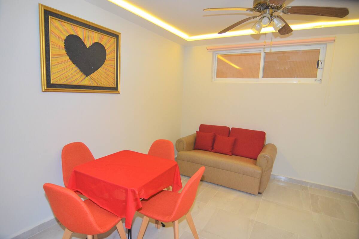 A dining area with a table and chairs, a sofa with red cushions, and a ceiling fan.