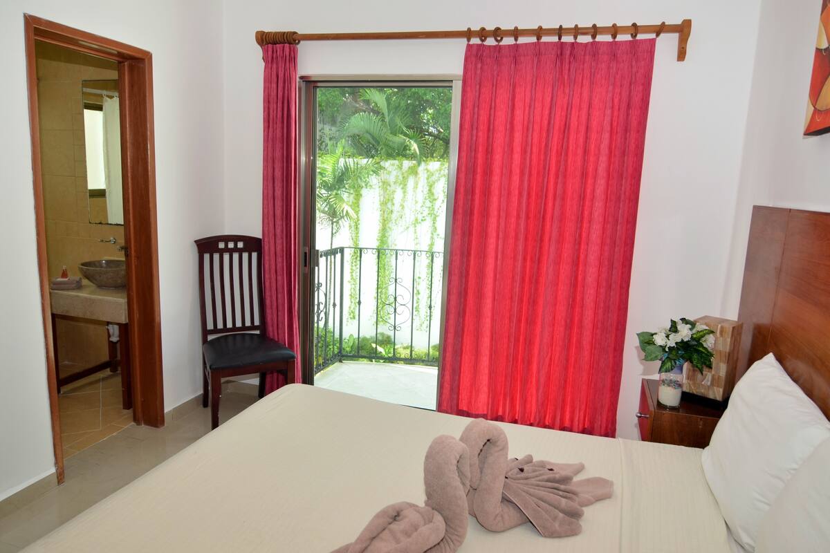 A hotel room with a bed, a chair, a small table with a vase, and a view of a balcony with red curtains.