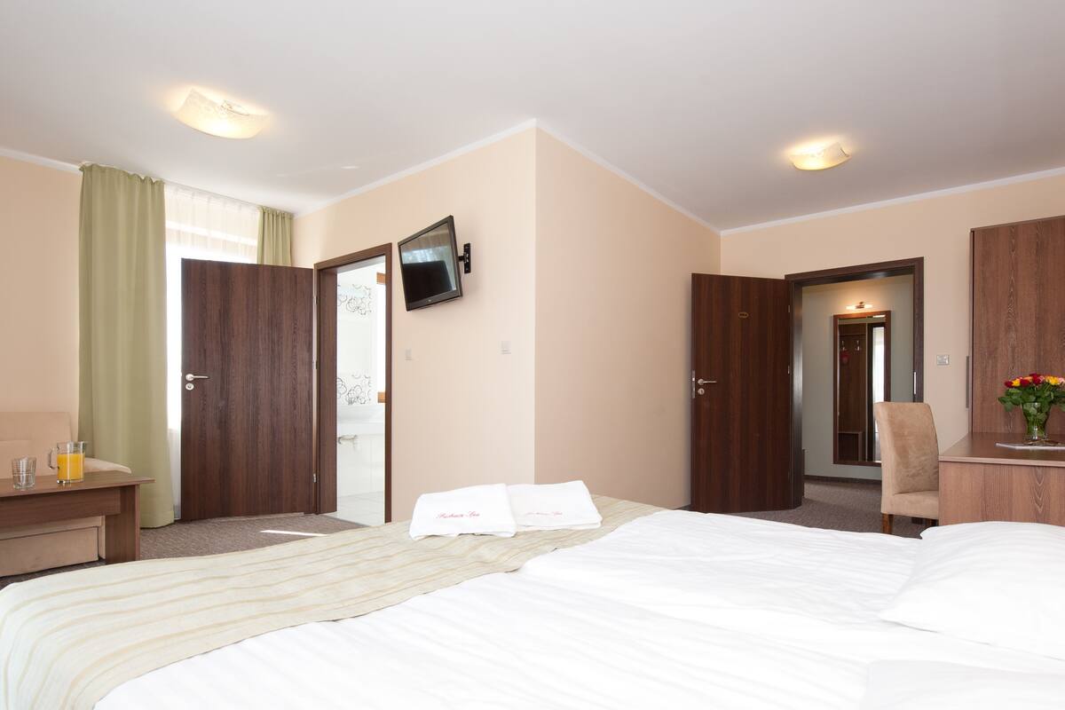 A hotel room with a bed, a desk with a lamp, a TV mounted on the wall, and a door leading to another room.