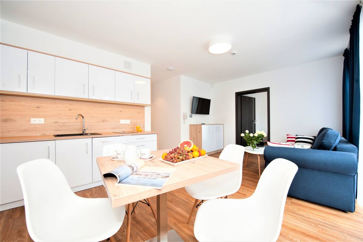 A modern living room with a blue sofa, a wooden dining table with chairs, and a kitchen area with white cabinets and a wooden countertop.