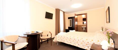 Habitación de hotel con cama, escritorio, silla, televisión y baño visible a través de una puerta abierta.
