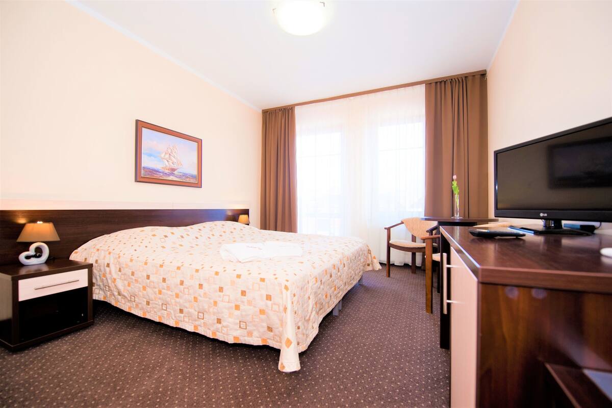 A hotel room with a large bed, a desk with a computer, a TV, a nightstand, and a window with curtains.