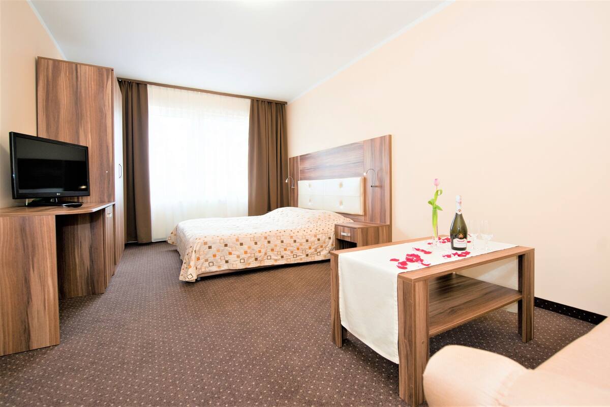 A hotel room with a bed, a TV, a bedside table with a bottle and glasses, and a chair.