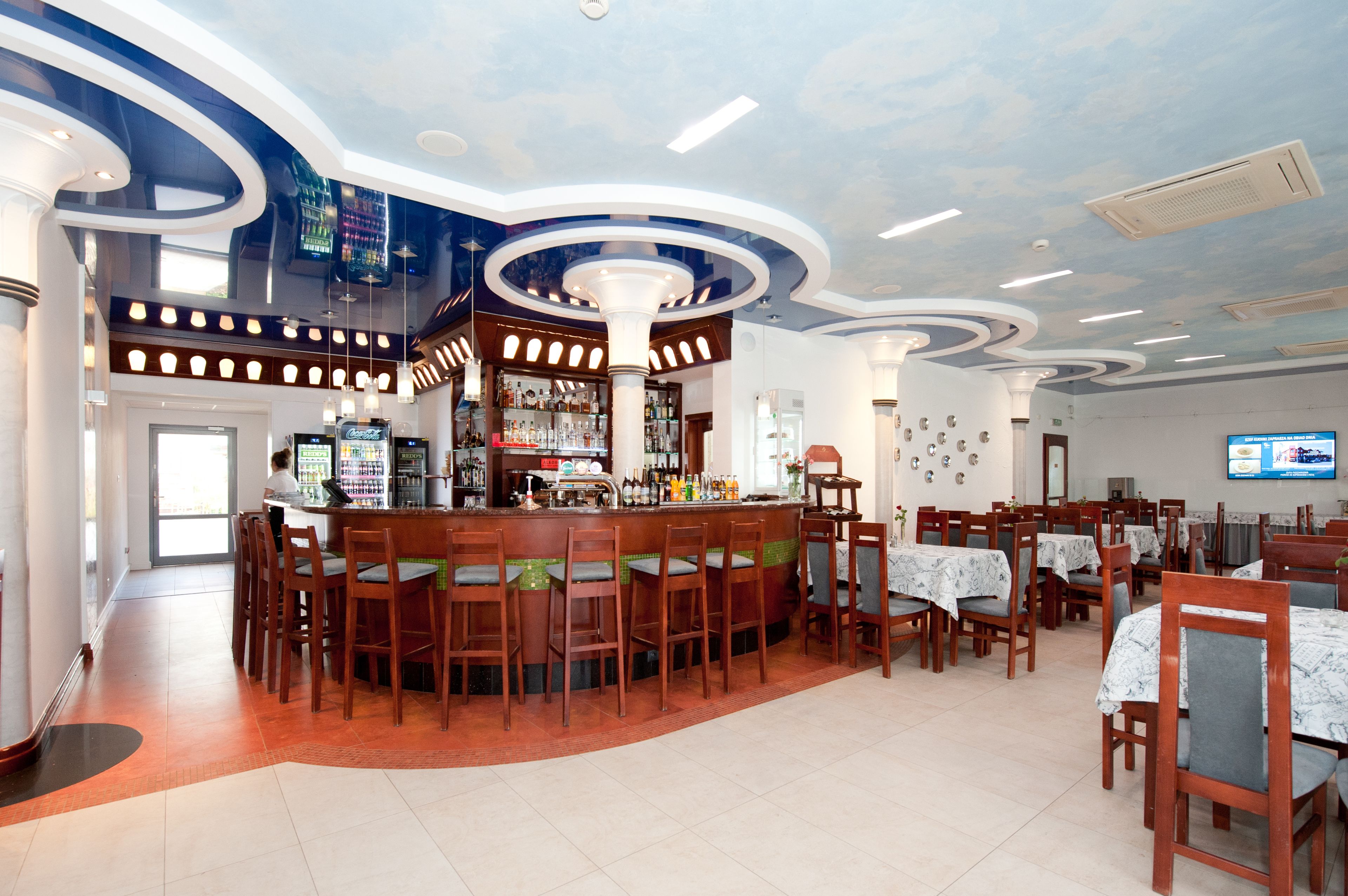 A restaurant with a bar area, multiple tables and chairs, and a ceiling with a unique design.