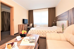 A hotel room with a bed, a desk with a computer, a chair, a coffee table with a cake and a magazine, and a vase with flowers. - Puchacz Spa (Rewal)