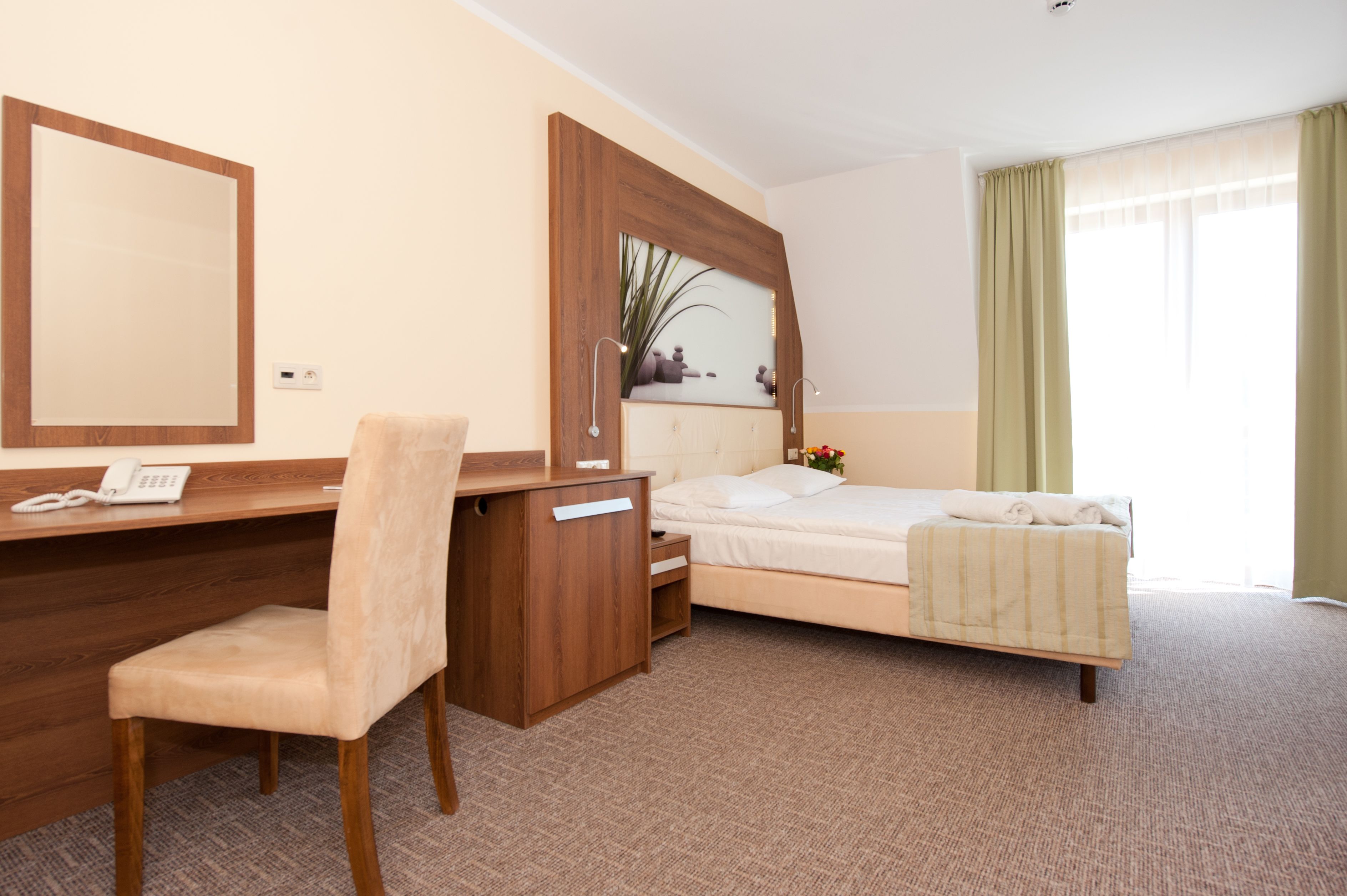 A hotel room with a wooden desk, a chair, a bed, a large mirror, and a window with curtains.