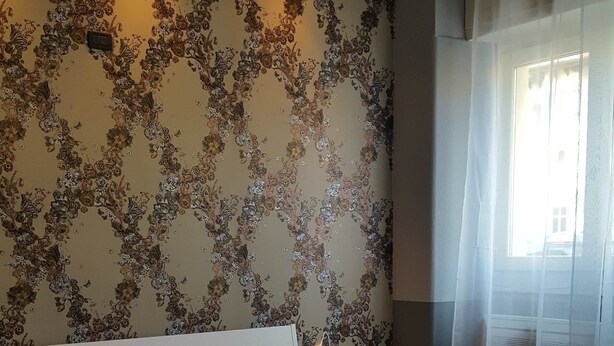 A neatly made bed with a white headboard, a bedside table with a lamp, and a floral-patterned wall. - Verona4rent Giulia (Verona)
