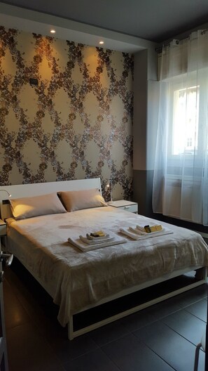 Cama bem arrumada com cabeceira branca, mesa de cabeceira com abajur e parede com padrão floral.