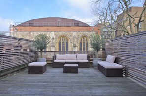 An outdoor seating area with a sofa and ottoman, a wooden deck, and a building with arched windows in the background.