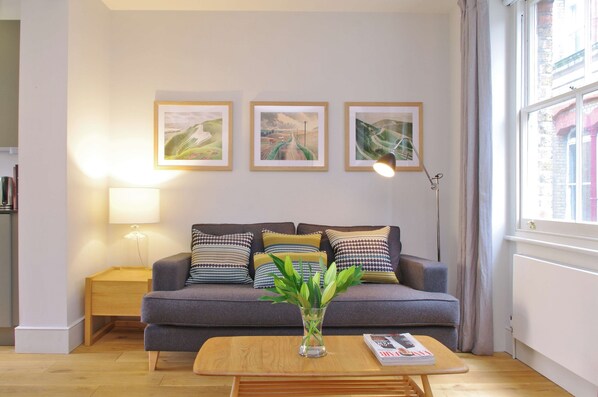 A living room with a grey sofa, a wooden coffee table, and a vase with green plants. There are three framed pictures on the wall, a floor lamp, and a window with curtains.
