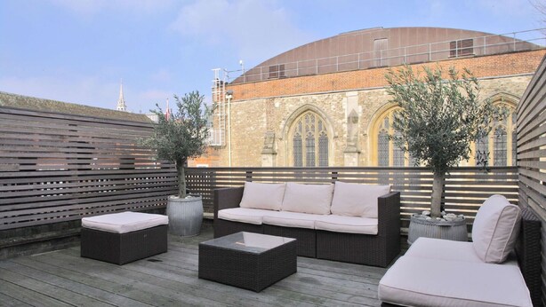 Eine Dachterrasse mit Korbmöbeln, einem Blumentopfbaum und Blick auf ein historisches Gebäude mit Rundbogenfenstern.