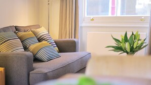 Ein graues Sofa mit gemusterten Kissen, eine Stehlampe und ein Topf mit Pflanze in einem Zimmer mit Fenster und Heizkörper.