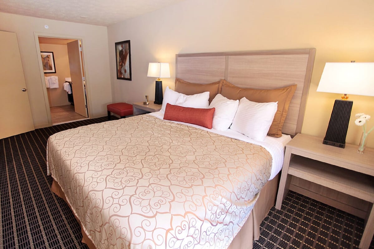 A hotel room with a large bed, two bedside tables, a nightstand, a door leading to a bathroom, and a small red bench.