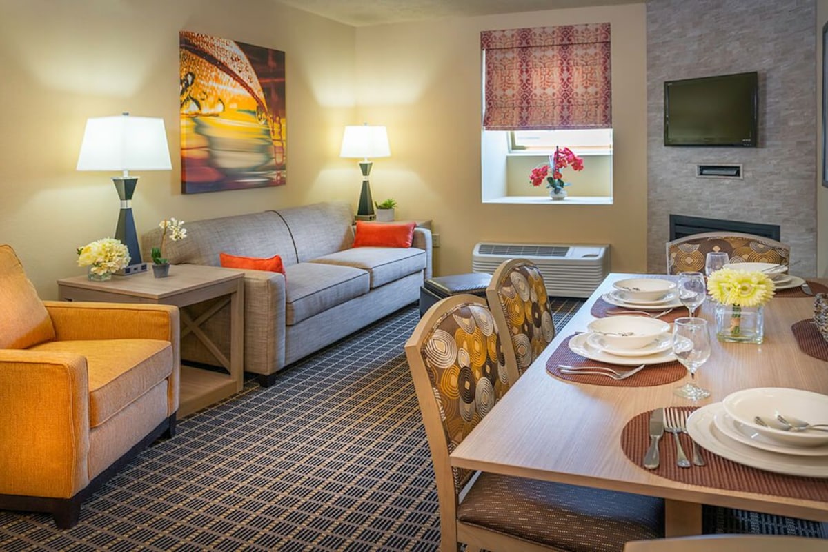 A hotel room with a sofa, armchair, dining table set for two, a TV mounted on the wall, and a window with a view.