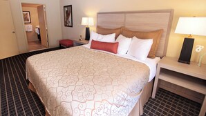 Quarto de hotel com uma cama grande, uma mesinha de cabeceira com abajur, uma pequena mesa com um vaso de flores e uma porta que dá para outro cômodo.