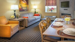 Quarto de hotel com sofá, poltrona, mesa de jantar posta para dois e TV na parede.