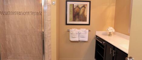 A bathroom with a shower, a framed artwork, a towel rack with towels, a vase with flowers, and dark cabinets.