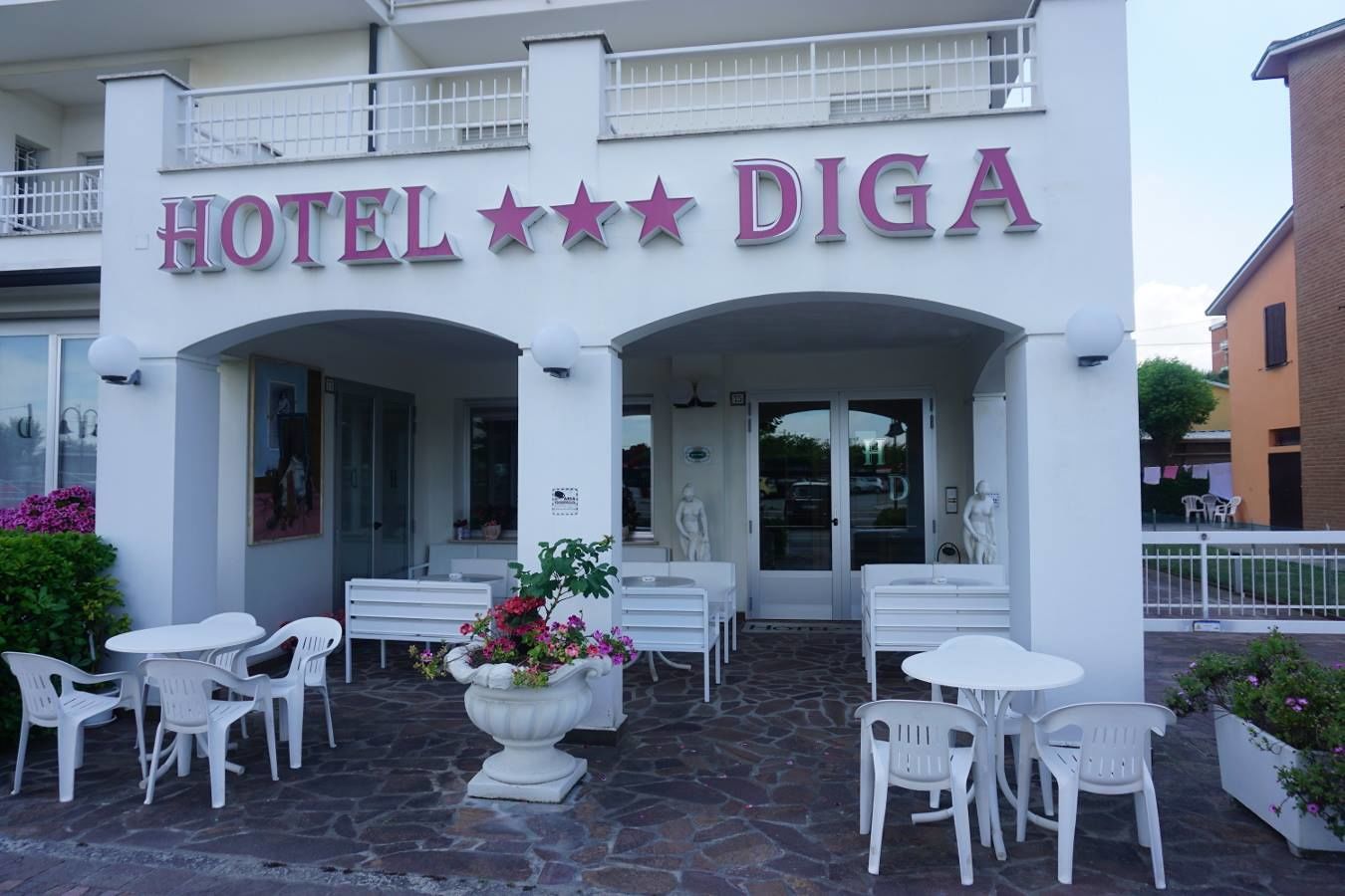 A hotel named HOTEL DIGA with a white facade, multiple seating areas with white plastic chairs, and a stone-paved entrance.