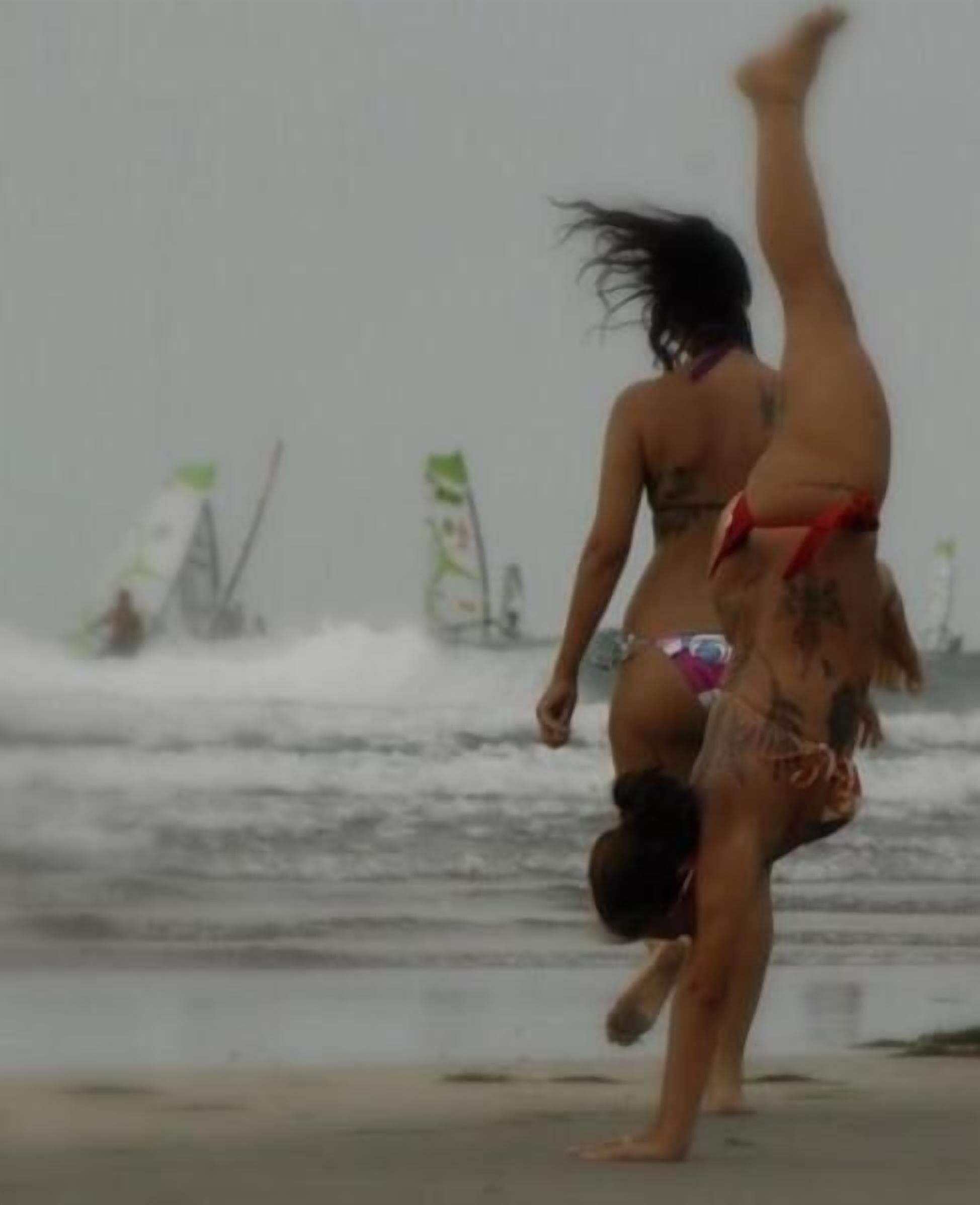 Escena de playa con dos personas haciendo un pino y tablas de surf al fondo.