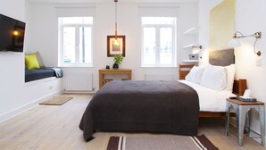 A modern bedroom with a large bed, a bedside table, a small desk, a chair, a TV mounted on the wall, and two windows with white curtains. - The London Agent Fitzrovia Studio 1 (London)