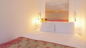 A bedroom with a bed, white bedding, a colorful throw blanket, a wall-mounted painting, and two wall-mounted lamps. - The London Agent Fitzrovia Studio 2 (London)